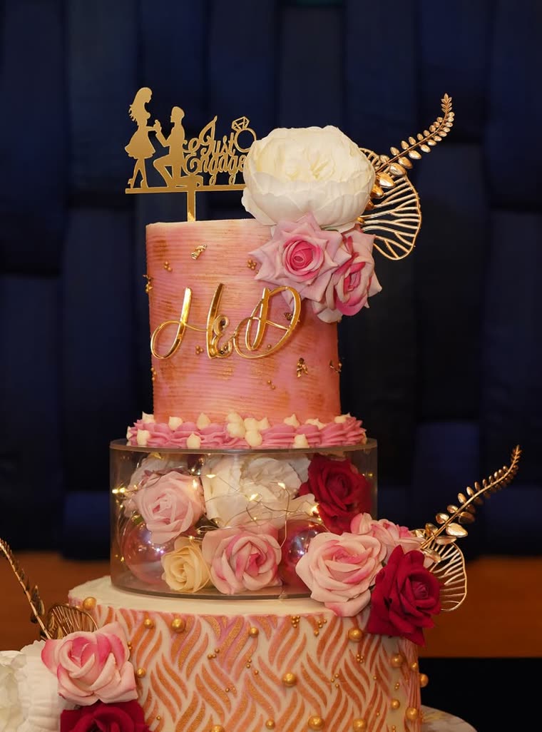 A three-tier engagement cake in blush and gold with fresh roses, a transparent floral tier lit from within, and a gold calligraphy topper.